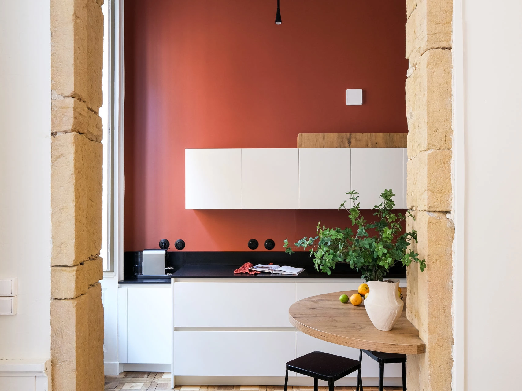 Cuisine au mur terracotta vue à travers un encadrement en pierre dorée, table ronde et tabourets, ESQUI STUDIO Lyon