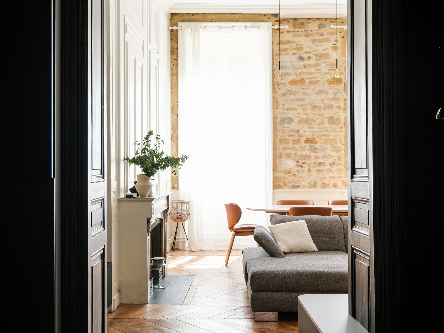 Perspective depuis une porte noire vers un séjour haussmannien : canapé gris, pierre apparente et cheminée, ESQUI STUDIO